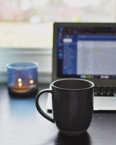 A photo of a gray coffee cup in focus at a desk. A laptop, burning candle, and window are out of focus.