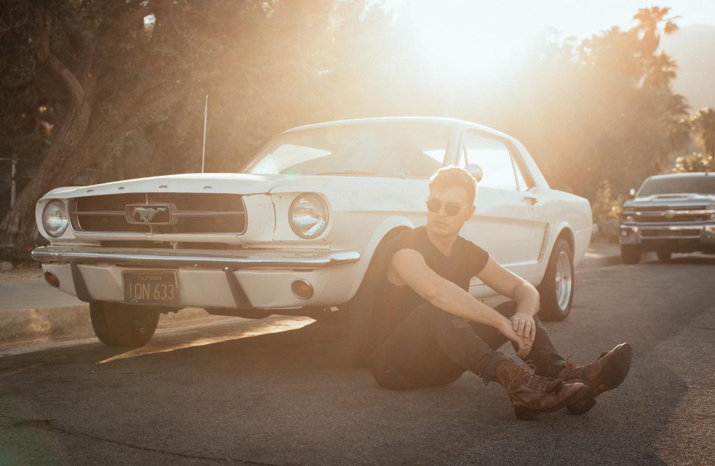 A stylized image of a man sitting on the ground beside an antique Mustang.